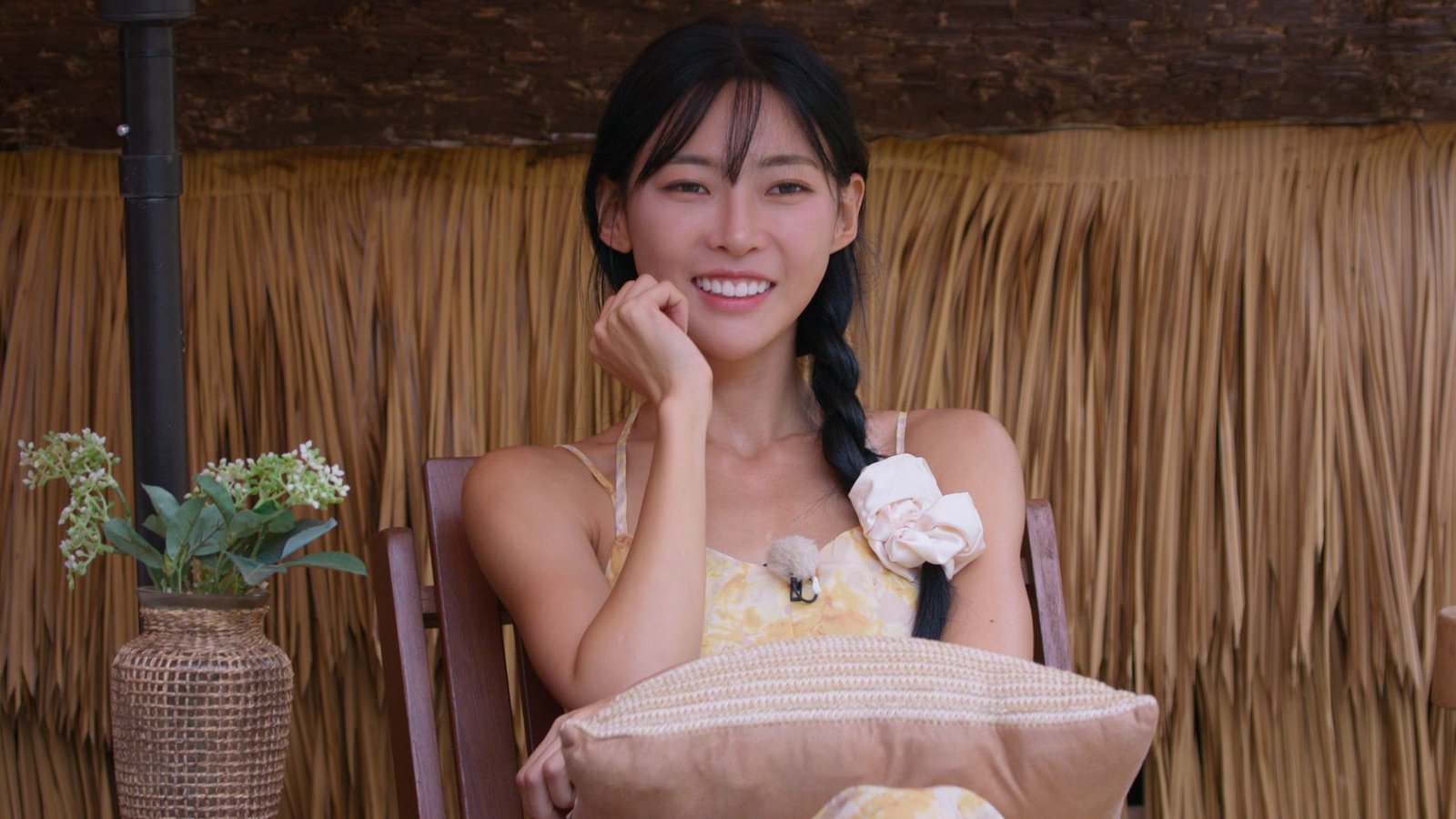 A woman (Kim Min-gee) wearing a yellow dress and a white scrunchie around her hair braid. She sits with a pillow on her lap, in front of a straw wall and next to white flowers in a vase, in the Netflix Korean reality show 'Single's Inferno' season 5.