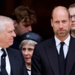 Prince William turns to Prince William and smiles at him, while both wear black suits with black ties and white shirts