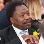 Professor Christopher Balogun-Lynch, sitting in a room, looking off to the right. He has a suit on, with a white shirt, blue tie, black jacket with a yellow rose on his lapel. He has short dark hair and has a moustache.