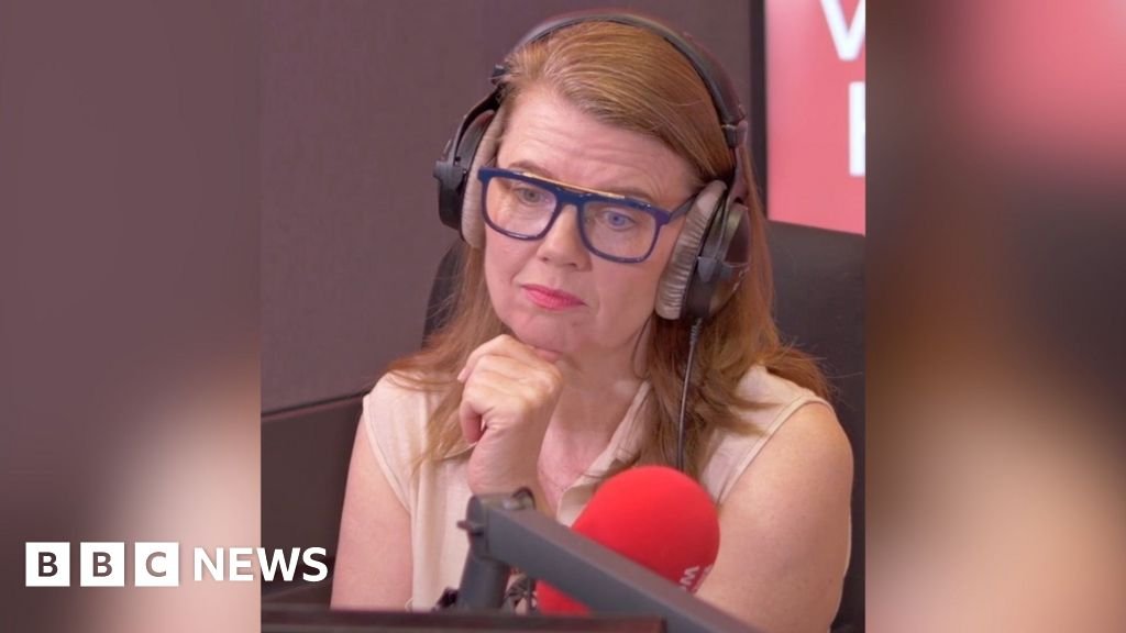 A woman with shoulder-length light ginger hair sits in front of a microphone and looks at someone off-screen